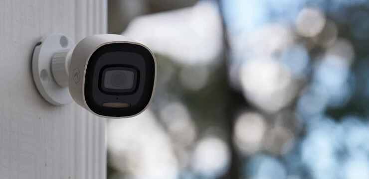 White ADT Smart Camera mounted outside a home in daylight, with garden foliage behind