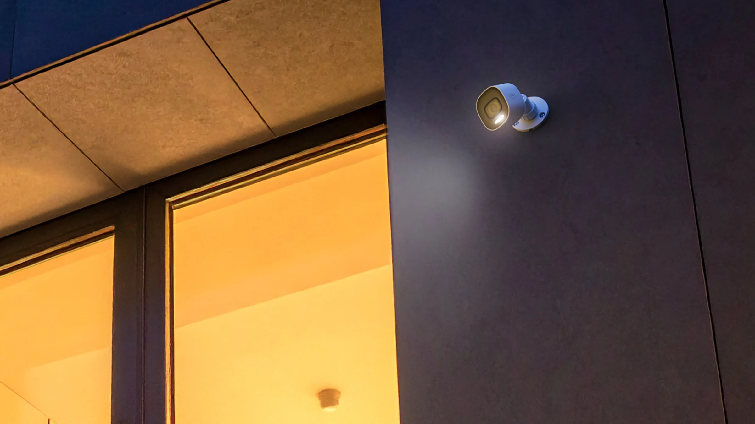 Outdoor Smart Camera with its spotlight switched on above warmly lit windows at dusk