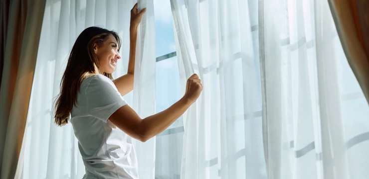 A woman opening the curtains beside a bright window at home